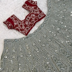 A flat lay of a light green lehenga skirt with silver embroidery and mirrors, and a dark red embroidered choli, on a white textured surface.