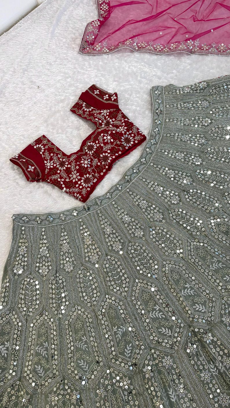 A flat lay of a light green lehenga skirt with silver embroidery and mirrors, and a dark red embroidered choli, on a white textured surface.