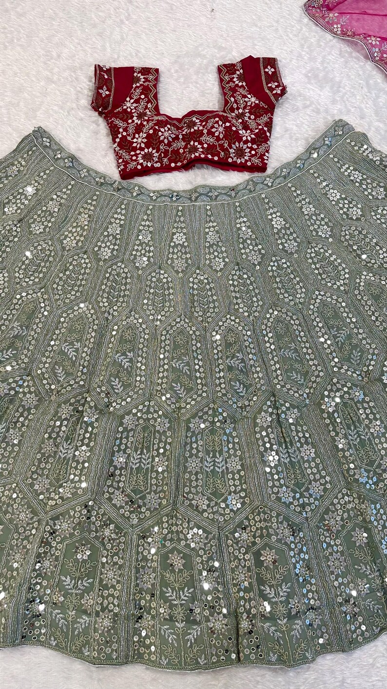 A flat lay of a dark red embroidered choli and a light green lehenga skirt with silver embroidery and mirrors, on a white textured surface.