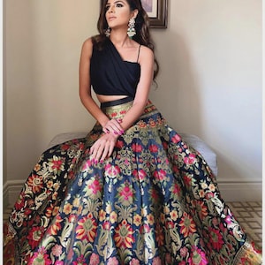 Peut inclure: Un haut court noir à une épaule associé à une jupe longue taille haute, dotée d'une base bleu foncé et d'un motif floral brocart complexe et multicolore dans des tons rose, or et vert, avec une large bordure métallique sur le bas.