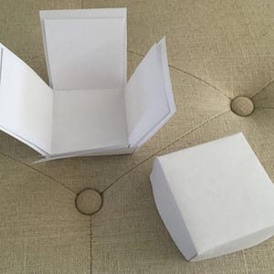 May include: Two white paper boxes, one open and one closed, are on a beige, tufted surface. The open box has its sides folded out, showing the inside. The closed box is a simple square shape. The background is a neutral colour.