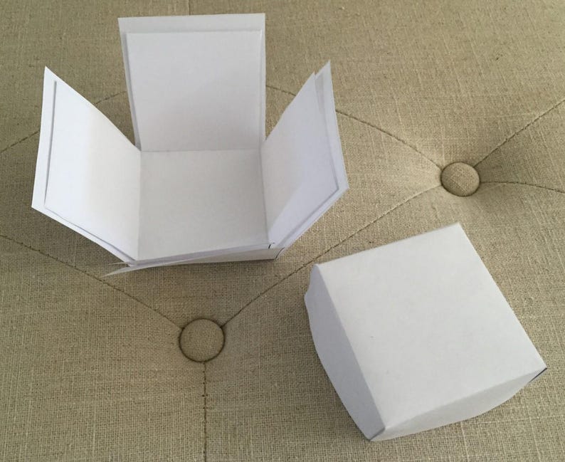 May include: Two white paper boxes, one open and one closed, are on a beige, tufted surface. The open box has its sides folded out, showing the inside. The closed box is a simple square shape. The background is a neutral colour.