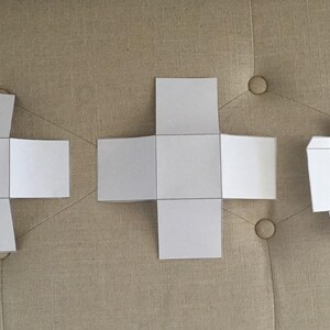 May include: Three white paper box templates with black outlines are displayed on a neutral-toned surface. The templates are in different stages of assembly, showcasing the folding process. The designs are simple and geometric.