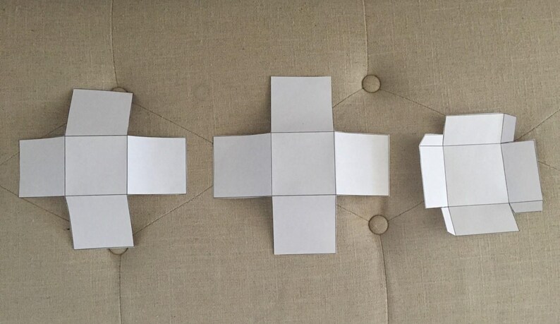 May include: Three white paper box templates with black outlines are displayed on a neutral-toned surface. The templates are in different stages of assembly, showcasing the folding process. The designs are simple and geometric.