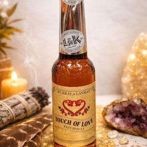 Puede incluir: Una botella de vidrio marrón de Florida Water Murray & Lanman, etiquetada como "Touch of Love Patchouli". La botella tiene una tapa negra y detalles plateados, con una etiqueta de estilo vintage con un diseño de corazón. El fondo incluye plumas y cristales.