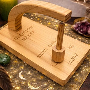 Puede incluir: Un "Decision Maker" de madera con un asa y una pequeña clavija. La base está grabada con "Decision Maker", "Never Always", "No" y "Maybe". Una vela verde encendida y una pluma están en el fondo.