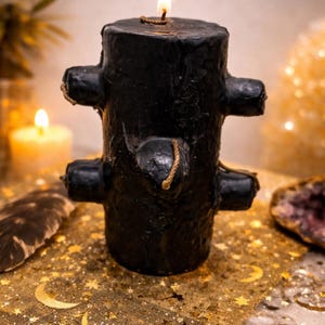 May include: A burning black candle with a distinctive, textured design and protruding features. The candle is placed on a gold cloth decorated with moon and star motifs. A lit candle and a feather are also visible in the image.