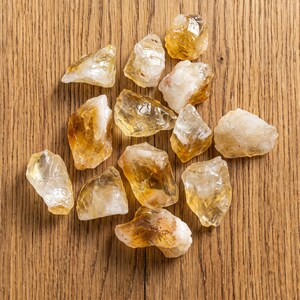May include: A collection of raw citrine crystals, showcasing a range of golden yellow and translucent hues. The crystals are irregularly shaped and vary in size, resting on a light brown wooden surface.