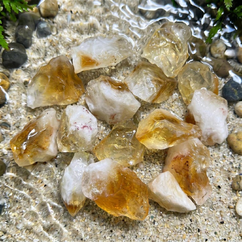 May include: A collection of raw citrine crystals, showcasing a range of golden yellow and white hues. The crystals are partially submerged in water, creating a shimmering effect. The image highlights the natural beauty and unique formations of the gemstones.
