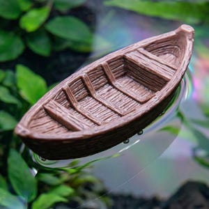Puede incluir: Un pequeño bote de madera marrón con tablones detallados, flotando sobre una superficie clara. El bote está rodeado de plantas acuáticas verdes, creando una escena natural. Este bote en miniatura es ideal para acuarios o terrarios.