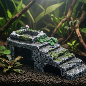May include: A gray and black aquarium decoration resembling a stone bridge with stairs and a small pool. Green moss and pink flowers add color to the textured surface. The bridge has an arched opening and is designed for aquatic environments.