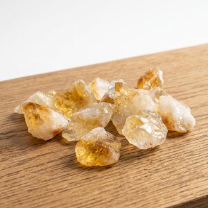 May include: A collection of raw citrine crystals, varying in size and shape, with a translucent, golden-yellow hue. The crystals are arranged on a light brown wooden surface, creating a natural and earthy aesthetic.