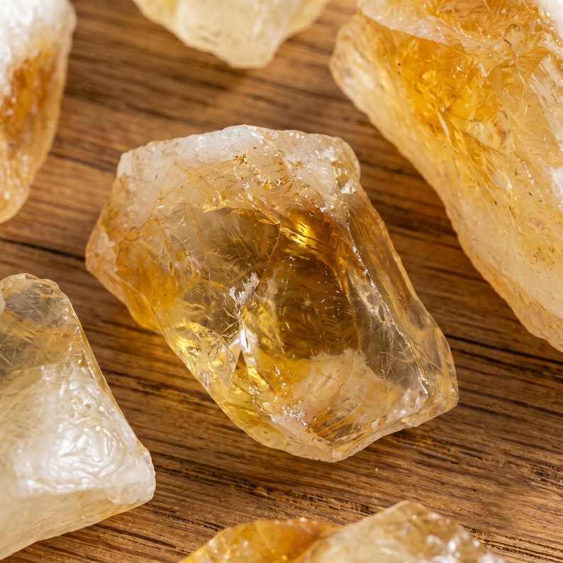 May include: Close-up of several raw citrine crystals, showcasing their natural, irregular shapes and translucent, golden-yellow hues. The crystals are scattered on a wooden surface, highlighting their varying sizes and textures. The image emphasizes the natural beauty of the gemstones.
