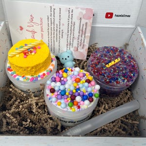 Op de afbeelding: Een geschenkdoos gevuld met drie bakjes kleurrijke slijm, waaronder een gele cake-vormige slijm met een kaars, een blauw berenfiguur en een doorzichtige spuitfles. De doos bevat ook versnipperd papier en een bedankbriefje.