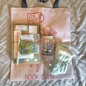 May include: A pink tote bag with black handles, featuring a red floral mug and open book design. The bag contains a stack of books, a glass jar with a floral design, a pair of light blue socks, and other small gifts. Text on the bag reads "BOOK CLUB".