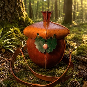 Puede incluir: Un bolso de cuero marrón con forma de bellota, con una tapa texturizada y una base lisa. El bolso presenta dos hojas de cuero verde sujetas al frente con un remache de color cobre e incluye una correa de hombro larga y ajustable de cuero.