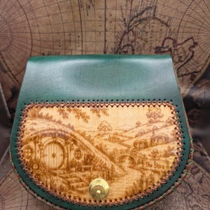 May include: A green leather pouch featuring a tan front panel with a detailed laser-engraved landscape of a hobbit-style home and rolling hills. The scene includes a round door, a stone bridge, and a sign that reads The Shire. A brass snap closure.