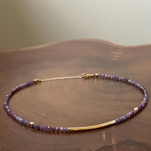 May include: A delicate necklace featuring small, faceted purple beads and gold accents. A gold bar is centered on the necklace. The necklace is displayed on a dark wooden surface.