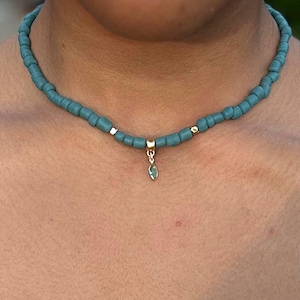 Könnte beinhalten: Eine Perlenkette mit kleinen, matten, blaugrünen zylindrischen Perlen, die aneinandergereiht sind. Die Kette enthält zwei kleine goldfarbene Zwischenperlen und einen zentralen goldfarbenen Anhängerhalter mit einem kleinen, baumelnden, marquiseförmigen blaugrünen Anhänger.