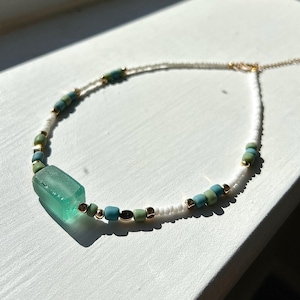 May include: A delicate necklace with a rectangular, seafoam green glass pendant. The necklace is strung with small white, teal, and green beads, with gold-coloured accents. The necklace is displayed on a white surface.