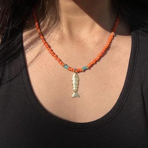 Puede incluir: Un collar con un colgante de pez en tono dorado suspendido de una hilera de pequeñas cuentas naranjas. El diseño incluye dos cuentas cilíndricas de color azul claro a los lados del colgante central, con pequeños espaciadores dorados en la cadena.