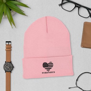 Op de afbeelding: Een lichtroze beanie met een zwart hartvormig Amerikaans vlagontwerp en het woord "FORENSICS" eronder. De beanie is gemaakt van een zacht, gebreid materiaal.