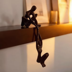 Puede incluir: Escultura de color bronce que representa dos figuras humanas, una sentada y otra colgando. La figura sentada extiende la mano hacia la figura colgante. La escultura está colocada sobre una estantería marrón oscuro.