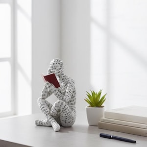 Peut inclure: Figurine blanche représentant une personne lisant un livre, recouverte de texte noir. La figurine est assise avec un livre rouge dans les mains. Une petite plante succulente en pot, des livres et un stylo sont sur la table.