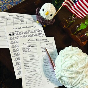 May include: A crochet planner, row counter, and organizer are displayed with a white yarn ball, a crochet hook, and a small crocheted eagle. An American flag and small glass bottles with greenery are also visible. The planner includes the text "Eagles".