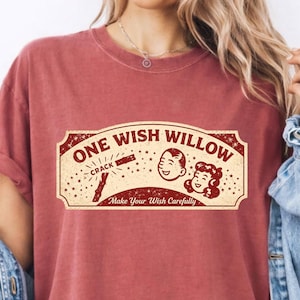 May include: A brick-red short-sleeve t-shirt featuring a cream-colored rectangular graphic with the text ONE WISH WILLOW, CRACK, and Make Your Wish Carefully. The graphic includes illustrations of a wishbone and two smiling faces.