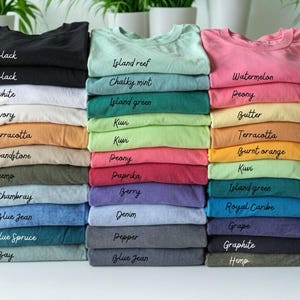 May include: Stacks of folded t-shirts in various colors, including black, white, and blue. Each stack is labeled with the color name in a handwritten font. The shirts are neatly arranged, showcasing a range of hues from pastels to darker shades.