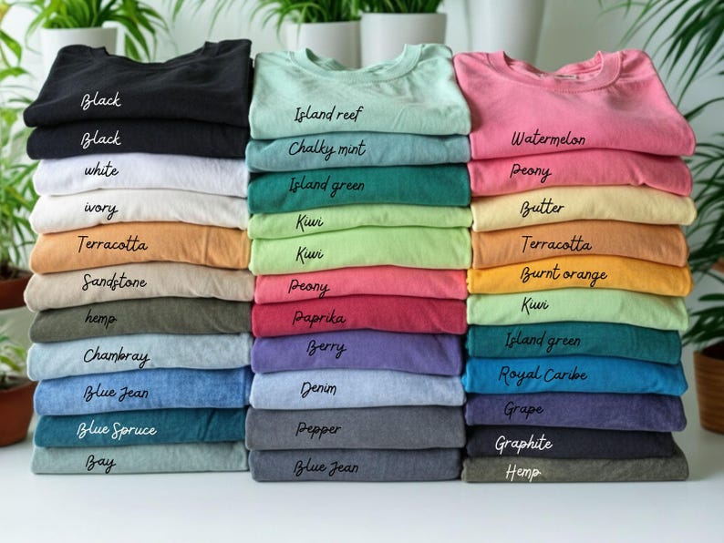 May include: Stacks of folded t-shirts in various colors, including black, white, and blue. Each stack is labeled with the color name in a handwritten font. The shirts are neatly arranged, showcasing a range of hues from pastels to darker shades.