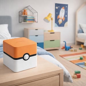Könnte beinhalten: Ein quadratisches, orangefarbenes, schwarzes und weißes Sparschwein mit einem kreisförmigen Knopfdetail. Die Spardose steht auf einem Nachttisch aus Holz in einem Kinderzimmer. Das Zimmer hat ein Bett, eine Kommode und einen bunten Teppich.