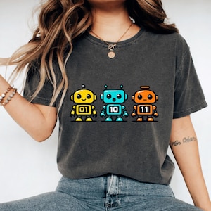 Könnte beinhalten: Dunkelgraues T-Shirt mit drei bunten Roboter-Grafiken. Die Roboter sind gelb, blau und orange, mit den Nummern 01, 10 und 11. Das Design befindet sich mittig auf dem Shirt.