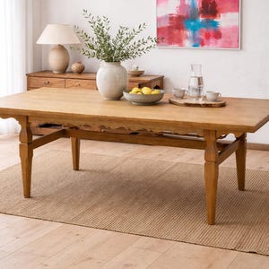 May include: A light brown wooden dining table with a decorative edge, set on a woven jute rug. The table is surrounded by a neutral-toned room with a window, a painting, and a potted plant.