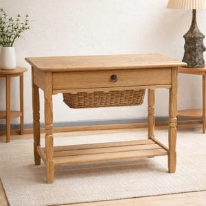 Puede incluir: Una consola de roble claro con un cajón, una cesta de mimbre y un estante de listones. La mesa tiene patas torneadas y un pomo oscuro en el cajón. La mesa mide 76 cm de largo.