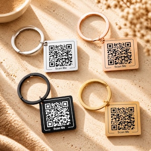 May include: Keychains with QR codes in silver, rose gold, gold, and black. Each keychain has a square tag with a QR code and the text "Scan Me". The keychains are on a sandy surface.