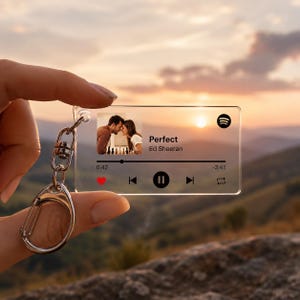Op de afbeelding: Een heldere acryl sleutelhanger met een muziekspeler interface met het nummer "Perfect" van Ed Sheeran. De sleutelhanger toont een foto van een stel en heeft een zilverkleurige metalen sluiting en ring. De achtergrond toont een zonsondergang.