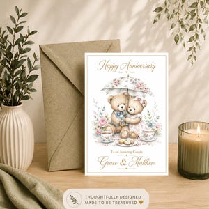 May include: A white anniversary greeting card featuring two illustrated teddy bears sitting under a floral umbrella. The text reads Happy Anniversary, To an Amazing Couple, and Grace & Matthew. The bears are surrounded by flowers and tea cups.