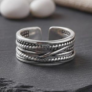 May include: A silver adjustable ring with a textured design. The ring has multiple bands with a braided and beaded pattern. The ring is open at the top and sits on a dark grey surface.