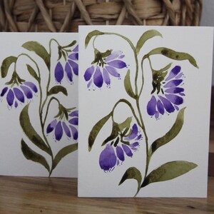 May include: Two greeting cards with watercolor floral designs. Each card features a sprig of purple bell-shaped flowers with green leaves and stems against a white background. The cards are displayed on a wooden surface.