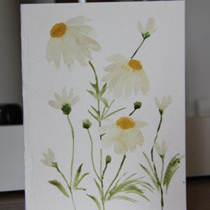 May include: A rectangular greeting card featuring a watercolor painting of white daisy flowers with yellow centers and green stems. The artwork displays several blooming flowers and small buds arranged on a textured white paper surface.
