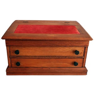May include: A wooden desk organizer featuring a rectangular frame with a hinged lid and two horizontal drawers. The top surface is inlaid with a rectangular red velvet panel. Each drawer is equipped with two small, dark, circular wooden knobs.