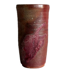 May include: A tall, cylindrical ceramic tumbler featuring a textured, mottled surface with deep red, brown, and earthy tones. The exterior displays a prominent, raised, and bubbly glaze patch in a vibrant magenta hue, contrasting with the matte finish.