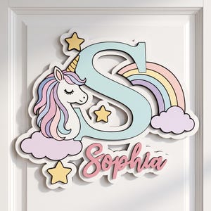 May include: A decorative wall sign featuring a large light blue letter S, a white unicorn with a pastel mane, a colourful rainbow, three yellow stars, and the name Sophia written in pink script, all mounted on a white layered backing.