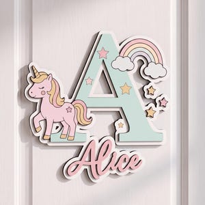 May include: A decorative wall sign featuring a large, light mint green letter A adorned with a pink unicorn, a pastel rainbow, and small stars. Below the letter, the name Alice is written in a cursive pink font. The entire piece has a white border.
