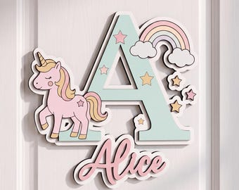 May include: A decorative wall sign featuring a large, light mint green letter A adorned with a pink unicorn, a pastel rainbow, and several small stars. Below the letter A, the name Alice is written in a cursive pink font with a white outline.