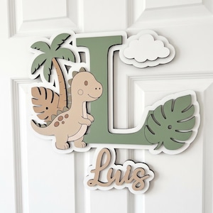 May include: A layered wooden wall sign featuring a large sage green letter L, a beige cartoon dinosaur, a palm tree, a monstera leaf, and a white cloud. Below the letter L, a separate wooden cutout displays the name Luis in cursive script.