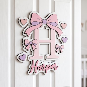 May include: A wall-mounted sign featuring a large, light pink letter H topped with a pink and purple bow. The sign is decorated with smaller pink and purple hearts and bows, and the name Harper is written in pink cursive script at the bottom.