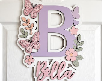 May include: A layered wall sign featuring a large light purple letter B and the name Bella in pink script. The design includes pink butterflies, small flowers, and green leaves on a white backing. The sign is mounted on a white paneled door.